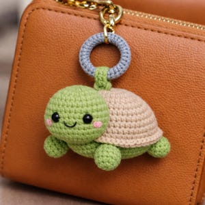 May include: A handmade crochet turtle keychain in green and tan, with a gray ring and gold-colored clasp. The turtle has a smiling face with pink cheeks, attached to a brown leather wallet.