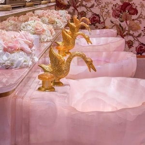 May include: A luxurious bathroom with pink onyx sinks and gold swan-shaped faucets. The sinks are arranged in a tiered design. Bouquets of white and pink flowers adorn the countertop. The wallpaper features a floral pattern.