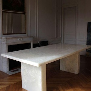 Könnte beinhalten: Ein großer, rechteckiger Tisch aus weißem Marmor mit zwei dicken, blockförmigen Stützen. Der Tisch steht auf einem Parkettboden. Der Raum hat einen Kamin und ein Gemälde an der Wand.