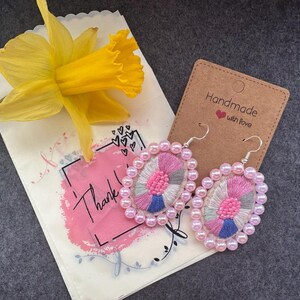 Könnte beinhalten: Ovale, bestickte Ohrringe mit rosa und grauem Faden, umgeben von rosa Perlen. Eine gelbe Narzisse und eine Karte mit der Aufschrift "Handmade with love" sind ebenfalls im Bild. Eine Geschenktüte mit dem Wort "Thank" ist ebenfalls im Bild.