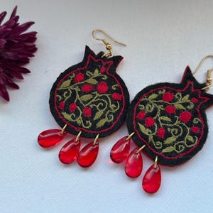 May include: Embroidered pomegranate earrings with red teardrop accents. The black felt earrings feature red and gold floral embroidery and gold-coloured ear wires. A dark purple flower is in the background.