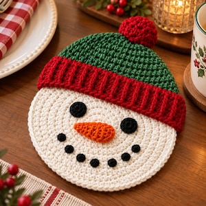 Puede incluir: Un posavasos redondo de ganchillo blanco con una cara de muñeco de nieve, ojos y boca negros circulares y una nariz naranja en forma de zanahoria. El muñeco lleva un gorro verde de ganchillo con borde rojo y un pompón rojo arriba.