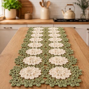 Pode incluir: Um caminho de mesa retangular com um padrão de crochê de crochê repetitivo de motivos florais em branco sujo, conectados por elementos de folhas em verde sálvia. O design intrincado de renda consiste em várias flores unidas em duas fileiras.