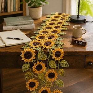 Può includere: Un lungo runner da tavola realizzato all'uncinetto a mano con un motivo ripetuto di girasoli gialli e marroni con foglie verdi. Il pezzo tessile floreale è drappeggiato su una superficie in legno, mostrando un intreccio di fili e petali.
