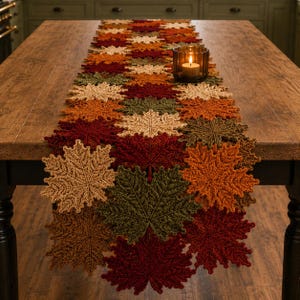 May include: A long table runner composed of interconnected crocheted maple leaves in shades of deep red, burnt orange, olive green, and cream. A small glass candle holder with a lit white pillar candle sits atop the runner on the wooden table.