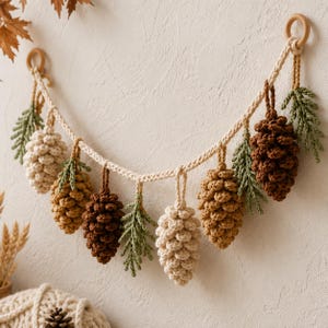 May include: A decorative garland featuring crocheted pinecones in shades of cream, tan, and dark brown, interspersed with crocheted green pine sprigs. The items are suspended from a thick, braided cream-coloured cord attached to two wooden rings.