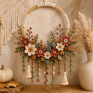 Op de afbeelding: Een ronde wandhanger van een houten borduurring met ingewikkelde gehaakte bloemen in roest-, crème- en blauwtinten, geaccentueerd met groen gehaakt loof en hangende crèmekleurige kwastjes, wat een gedetailleerd bloemstuk vormt.