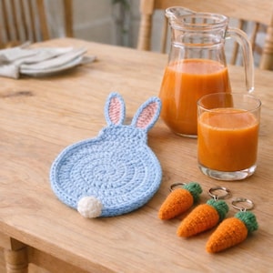 Może przedstawiać: Jasnoniebieski szydełkowy podkładka w kształcie królika z różowymi uszami wewnętrznymi i białym pomponem. Trzy pomarańczowe breloki w kształcie marchewki z zielonymi wierzchołkami leżą obok podkładki. Szklanka i dzbanek soku pomarańczowego stoją na drewnianym stole.