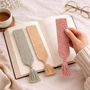 Könnte beinhalten: Drei handgefertigte Häkel-Lesezeichen in Grün-, Beige- und Rosatönen, jedes mit einem passenden Quastenanhänger. Ein Lesezeichen wird in einem Buch offen gehalten. Die Lesezeichen liegen auf einer weißen Oberfläche mit einer Tasse Tee und einer Brille im Hintergrund.