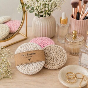 May include: Round, crocheted face scrubbies in cream and pink colors. One cream scrubby is wrapped in a brown paper band labeled "face scrubby Handmade" with a heart icon. The set includes textured, circular cotton pads for facial cleansing.