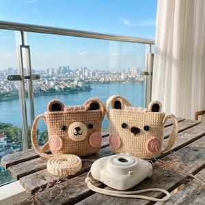 May include: Two crocheted bear-shaped bags, one in light brown and one in cream, each featuring dark brown ears, black button eyes, a stitched nose, and pink circular cheeks. A white instant camera with a strap sits in front of the bags.
