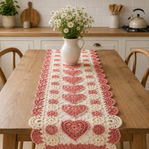 May include: A rectangular crochet table runner featuring a repeating pattern of dusty rose hearts and cream-colored circular motifs. The runner has a scalloped edge and is centered on a wooden table, topped with a white ceramic vase of daisies.