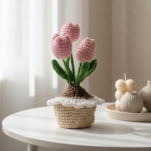 May include: A crocheted tulip arrangement in a beige pot. The arrangement features three pink tulips with green stems and leaves. The pot is beige with a white scalloped trim. The arrangement sits on a white table.