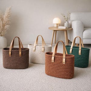 May include: Four crochet tote bags in shades of brown, beige, and green. Each bag has a rectangular shape with leather handles attached by leather straps. The bags are displayed on a neutral-colored carpet.