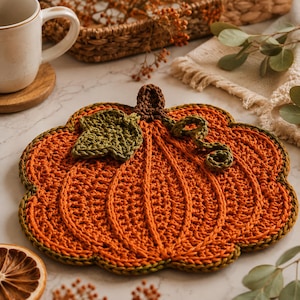 Pode incluir: Um descanso de panela em crochê com formato de abóbora, feito de fio laranja vibrante com um caule castanho escuro, uma folha verde texturizada e uma gavinhas verde. As bordas da abóbora são finalizadas com um contorno verde escuro e canelado.