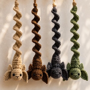 May include: Four hanging crochet ornaments in beige, brown, dark gray, and green. Each ornament features a spiral-shaped body attached to a round head with two large ears, black bead eyes, and a stitched mouth, suspended by a tan cord loop.