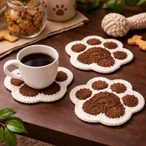 May include: A white ceramic coffee cup filled with dark coffee sits on a brown and white paw-shaped crochet coaster. Two additional paw-shaped coasters are visible. The image also includes a jar of dog treats and a dog toy.