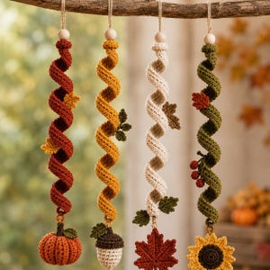 May include: Four hanging spiral ornaments made of crocheted yarn in autumn colours. Each piece features a wooden bead at the top, a twisted central column, and a bottom charm: a pumpkin, an acorn, a maple leaf, and a sunflower with berries.