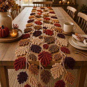 May include: A long, crocheted table runner featuring a repeating pattern of oak leaves in shades of cream, tan, orange, rust, and deep burgundy. The leaves are connected by thin, braided yarn stems that drape over the edge of the wooden table.