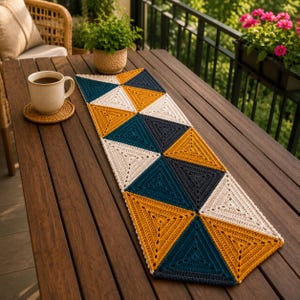 May include: A rectangular table runner made of crocheted triangles in mustard yellow, teal, dark navy blue, and cream. The runner features a geometric pattern of alternating colors and is laid flat on a dark brown wooden table surface.