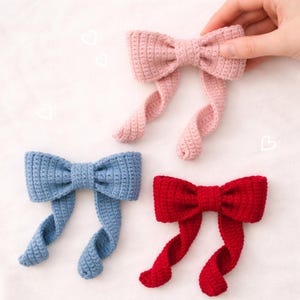 Puede incluir: Tres lazos de ganchillo hechos a mano en rosa, azul y rojo. Los lazos tienen una forma de lazo clásica con colas largas y rizadas. Una mano sostiene el lazo rosa. Los lazos están sobre un fondo blanco con pequeños garabatos de corazón.