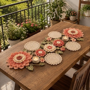 Peut inclure: Un chemin de table en crochet au motif floral composé de grandes fleurs superposées corail et crème, de petits napperons circulaires crème et de feuilles en crochet vertes. Deux bougeoirs gris avec des bougies chauffe-plat sont posés dessus.