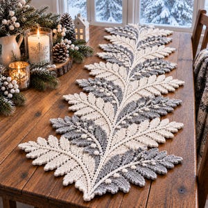 Può includere: Un lungo runner da tavola decorativo caratterizzato da un intricato motivo a foglie di felce lavorato all'uncinetto. Il design alterna filato color crema e grigio, con piccoli dettagli in rilievo lungo lo stelo centrale, creando texture.