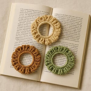 Puede incluir: Tres coleteros de ganchillo hechos a mano en beige, marrón y verde, colocados sobre un libro abierto. Los coleteros tienen un borde con volantes y un diseño circular, mostrando un estilo rústico y bohemio.