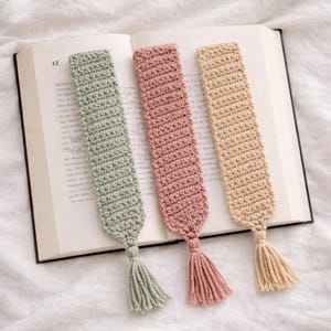 May include: Three handmade crochet bookmarks in pastel shades: sage green, dusky pink, and cream. Each bookmark has a matching tassel. The bookmarks are on an open book.