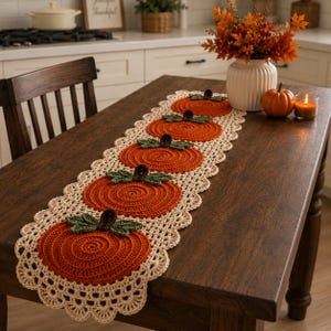 Puede incluir: Un camino de mesa de ganchillo largo y rectangular con cinco motivos circulares de calabaza naranja, tallos marrón oscuro y hojas verdes, bordeado por un encaje festoneado de color crema, extendido sobre una mesa de madera oscura.