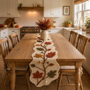 May include: A cream-coloured crocheted table runner with a scalloped edge, featuring an intricate vine pattern adorned with three-dimensional knitted autumn leaves in shades of rust, orange, olive green, and brown, along with small acorn accents.