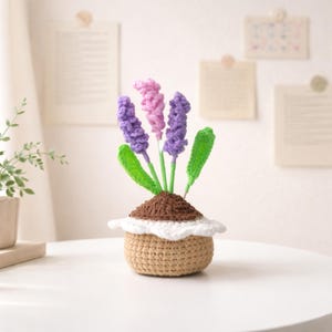 May include: A crocheted lavender plant in a beige pot. The plant features purple and pink flower heads, green leaves, and a brown soil base. The pot has a white, scalloped trim.