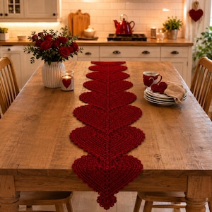 May include: A dark red crochet table runner featuring a repeating heart-shaped pattern laid out on a wooden table. The runner is centered on the table, which also holds a white vase filled with red roses and a stack of white plates with a mug.