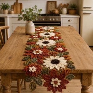Puede incluir: Un camino de mesa de ganchillo largo y rectangular con un patrón repetitivo de grandes girasoles blancos y marrón oscuro, flores más pequeñas de color óxido y hojas verdes texturizadas. El diseño floral está hecho de hilo.