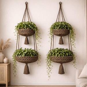 May include: Two hanging macrame plant holders with two tiers each, filled with green plants. The brown holders have a woven design and a tassel at the bottom. The plants have long, trailing vines. The holders are suspended from the ceiling.