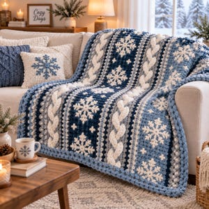 May include: A chunky knit throw blanket featuring a repeating pattern of white snowflakes on a dark blue and light blue background, accented with thick white cable knit stripes. The blanket is draped over a sofa with matching throw pillows.
