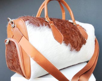 May include: A duffel bag with a cowhide pattern in white and brown. The bag has brown leather handles, accents, and a shoulder strap. A zippered pocket is on the side.