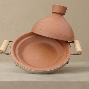 Peut inclure: Un plat à tajine en terre cuite avec un couvercle conique, posé sur une base métallique avec des poignées en bois. Le plat ouvert révèle son intérieur. Le tajine est un plat de cuisson traditionnel.