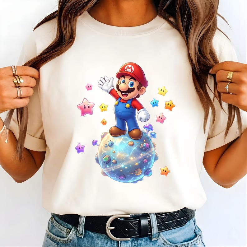 Peut inclure: T-shirt cr&egrave;me avec une illustration de dessin anim&eacute; de Mario faisant signe. Mario porte un chapeau rouge, une salopette bleue et des chaussures marron, debout sur une plan&egrave;te entour&eacute;e d'&eacute;toiles color&eacute;es. Le design est vif et ludique.