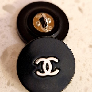 May include: Two black buttons with a silver-colored metal shank. One button features a white interlocking logo on its face. The buttons are round and appear to be made of a glossy material.