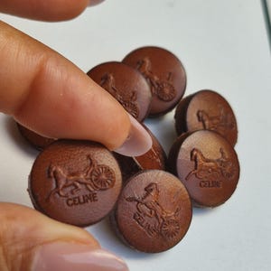 Könnte beinhalten: Eine Sammlung kleiner, runder, brauner Lederknöpfe, die jeweils mit einem Pferde- und Kutschenlogo sowie dem Schriftzug CELINE geprägt sind. Die Knöpfe haben eine glatte, kreisförmige Form und einen einheitlichen dunkelbraunen Farbton.