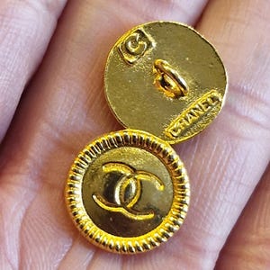 Peut inclure: Deux boutons ronds dorés présentant un logo CC entrelacé en relief sur le devant et une bordure texturée style corde. Le dos des boutons affiche le texte CHANEL et un petit poinçon carré avec une lettre G en son centre.
