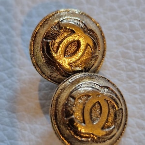 Peut inclure: Deux boutons ronds dorés avec une bordure texturée et un motif central de deux C entrelacés. Les boutons ont un aspect vintage ou antique, avec une finition légèrement usée. Les boutons sont probablement destinés aux vêtements ou à des fins décoratives.