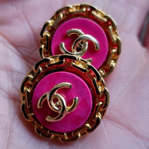 Könnte beinhalten: Ein Paar runde Ohrringe mit einem goldfarbenen Kettenglied-Rand. Jeder Ohrring hat eine leuchtend pinkfarbene Mitte mit einem verschlungenen goldenen Logo. Ein klassisches Accessoire.