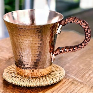 May include: A copper mug with a hammered texture and a woven handle. The mug sits on a round, woven coaster. The mug has a wide mouth and a copper-coloured interior. The handle is a dark brown colour.