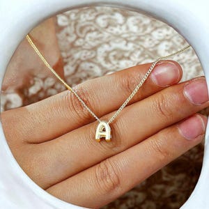 Könnte beinhalten: Eine goldfarbene Halskette mit einem Anhänger in Form des Buchstabens "A". Der Anhänger ist ein Großbuchstabe "A" in einer Serifenschrift. Die Halskette hat eine zarte, dünne Kette. Die Halskette wird auf einer Hand präsentiert.
