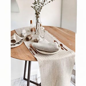 Puede incluir: Una mesa redonda de madera preparada para cenar con un camino de mesa de lino de tonos neutros. La mesa está puesta con platos, servilletas, tenedores, cucharas y botellas decorativas. Un jarrón de vegetación añade un toque natural.