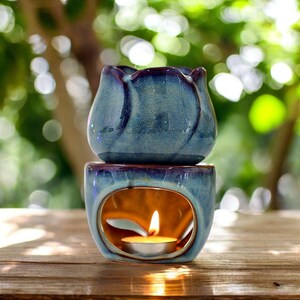 May include: A two-piece ceramic essential oil diffuser in shades of blue with a glossy finish. The top piece has a flower-like shape. The bottom piece has an oval opening with a lit tea light candle inside, creating a warm glow.