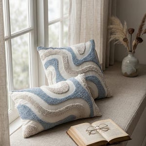 Könnte beinhalten: Zwei dekorative Kissen mit einem wellenförmigen Muster in Hellblau, Beige und Weiß. Die Kissen liegen auf einer Fensterbank neben einem offenen Buch und einer Brille. Eine Vase mit getrockneten Pflanzen befindet sich im Hintergrund.