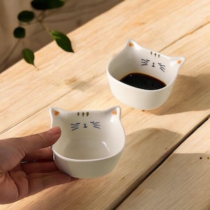 May include: Two white ceramic cat-shaped bowls with black facial details and orange ear accents. One bowl contains a dark liquid. The bowls are on a wooden surface. The bowls are designed for serving sauces or dips.